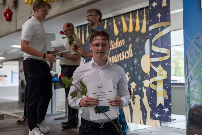 Ein junger Mann mit Brille nimmt eine rote Rose und eine Mappe mit der Aufschrift „Zeugnismappe ESBK Heller“ entgegen. Im Hintergrund sind weitere Personen und eine Dekoration mit der Aufschrift „Glücklichen Wunsch“ zu sehen.