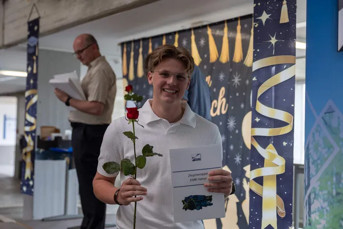 Junger Mann mit roter Rose und Zeugnismappe lächelt in die Kamera. Im Hintergrund eine festliche Dekoration mit silbernen Girlanden und Sternen.