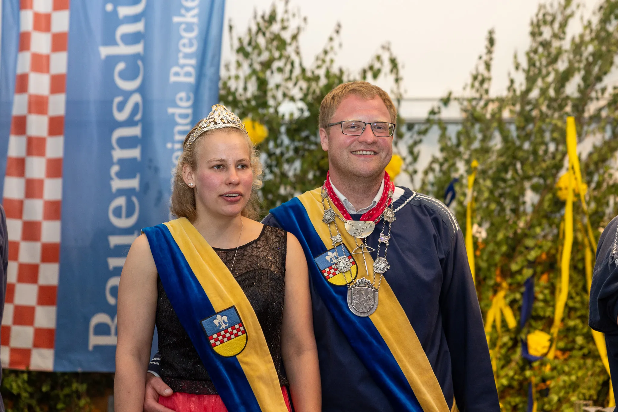 Eine junge Königin mit Tiara und ein Mann mit Kette und blauem Band stehen lächelnd vor einer rot-weiß karierten Fläche. Im Hintergrund ist ein Festzelt mit grünen Pflanzen zu sehen.
