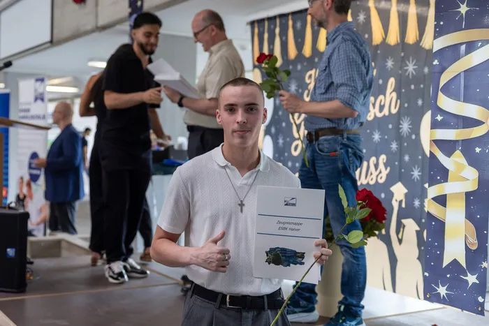 Junger Mann mit kurzem Haar hält eine Zeugnismappe und eine rote Rose. Im Hintergrund sind weitere Personen und eine Dekoration mit dem Schriftzug 