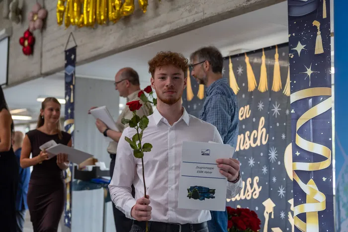 Junger Mann mit roten Haaren hält eine einzelne rote Rose und eine Zeugnismappe in der Hand. Im Hintergrund sind weitere Personen und festliche Dekorationen zu sehen.