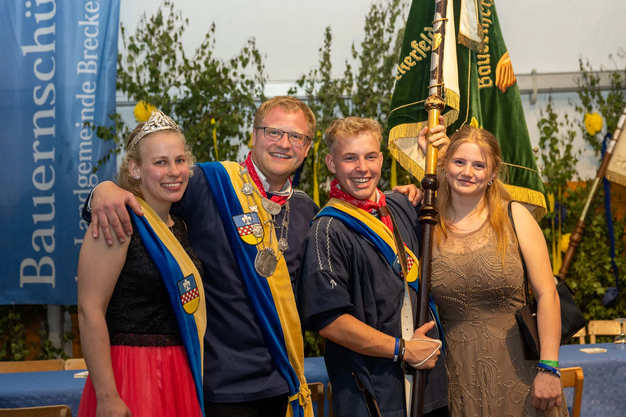 Drei junge Frauen und ein Mann in traditioneller Tracht posieren lächelnd für ein Foto. Der Mann trägt eine goldene Kette und hält eine Flagge.