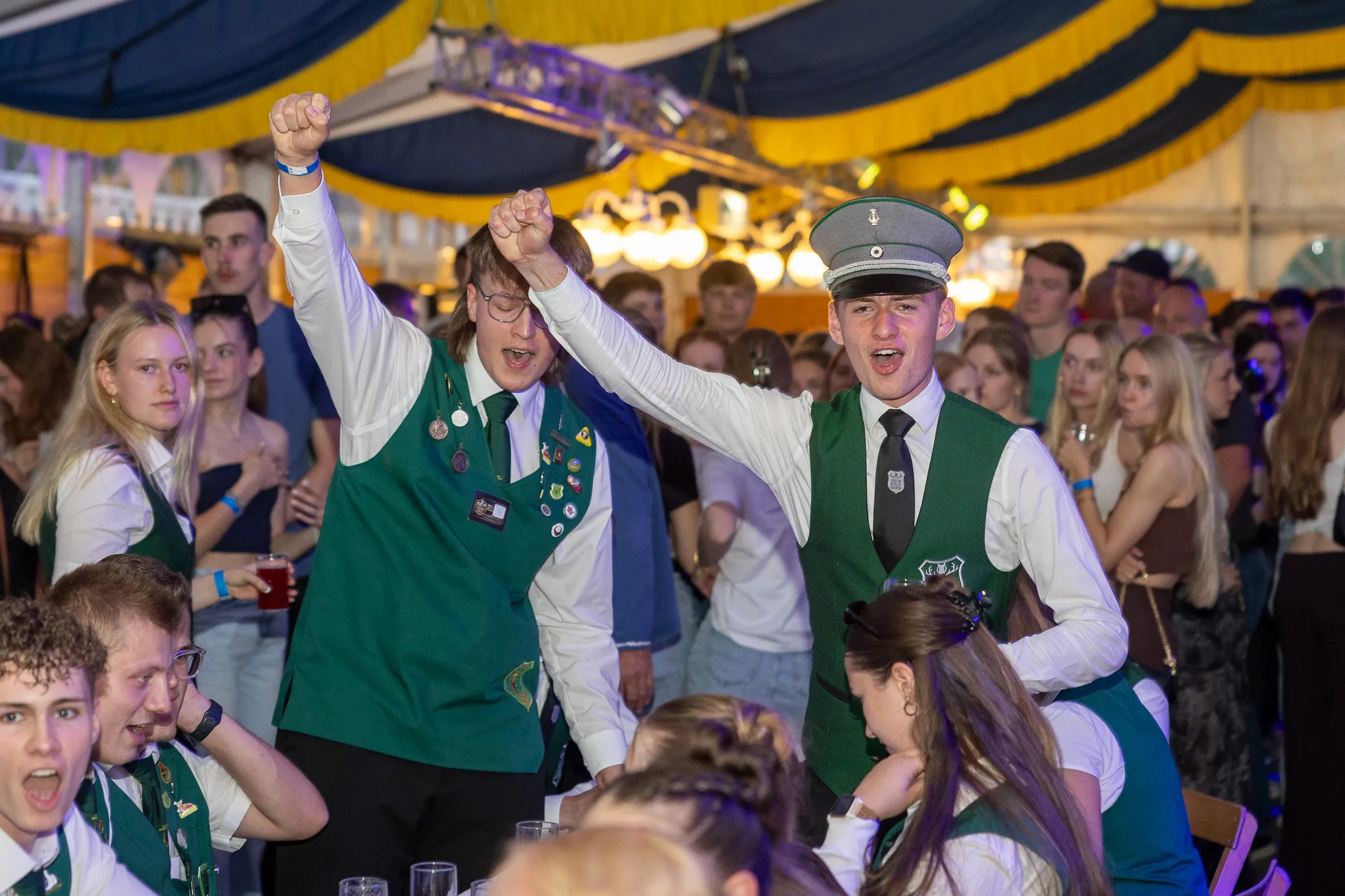Zwei junge Männer in grünen Trachtenwesten und ein Mann in Uniform stehen im Fokus eines belebten Festzeltes, während sie enthusiastisch in die Luft jubeln.