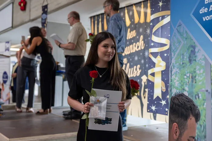 A young woman in a dark dress smiles at the camera while holding a red rose and a certificate. In the background, a banner with golden lettering and other people are visible.