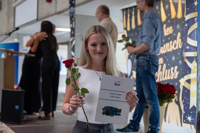 Junge Frau mit roter Rose und Zeugnismappe der ESBK Halver lächelt in die Kamera. Im Hintergrund sind weitere Personen und eine festliche Dekoration zu sehen.