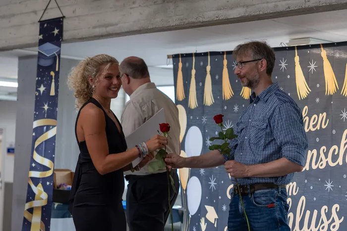Ein Mann übergibt einer Frau eine rote Rose bei einer Abschlussfeier der Höheren Handelsschule. Im Hintergrund ist ein Banner mit der Aufschrift „hen nsch m chluss“ zu sehen.