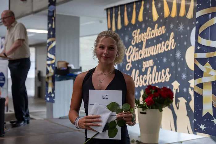 Junge Frau mit Diplom und Rose lächelt in die Kamera. Im Hintergrund ein Banner mit der Aufschrift „Herzlichen Glückwunsch zum Abschluss“.