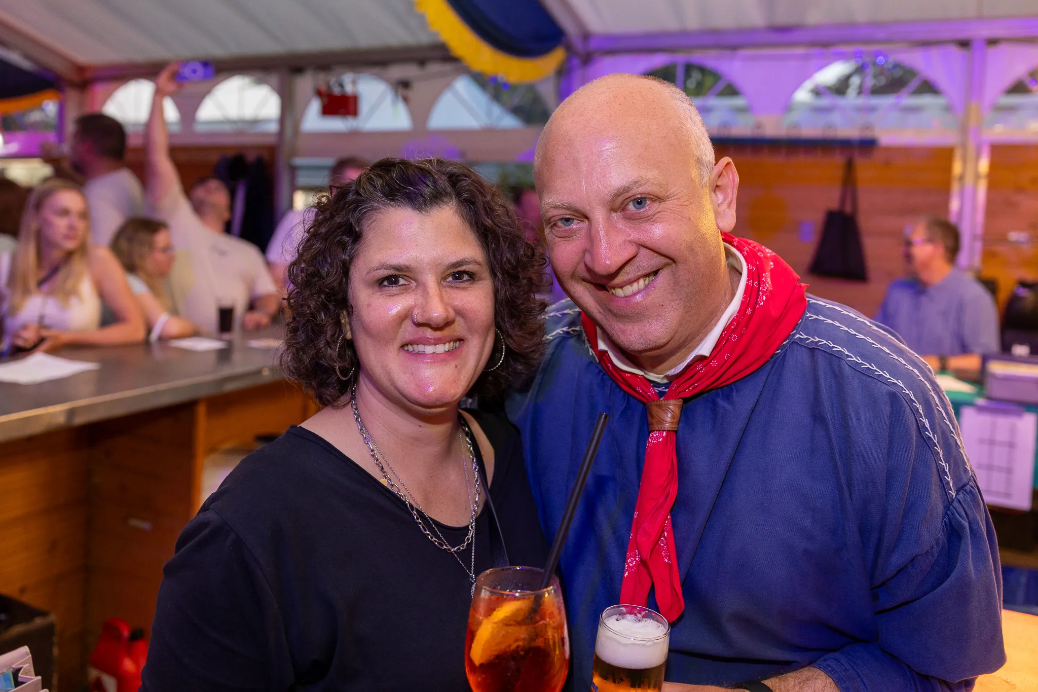 Zwei lächelnde Menschen, eine Frau mit lockigem Haar und ein Mann mit rotem Halstuch, posieren eng zusammen vor einer Bar in einem belebten Festzelt.