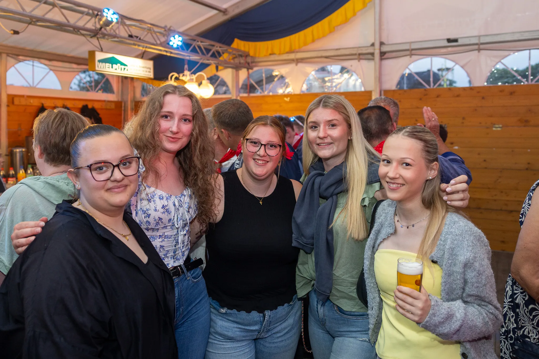 Fünf junge Frauen umarmen sich lächelnd im überfüllten Festzelt, eine von ihnen hält ein Bierglas in der Hand. Im Hintergrund sind weitere Menschen und ein Schild mit der Aufschrift „Wielpütz“ zu sehen.