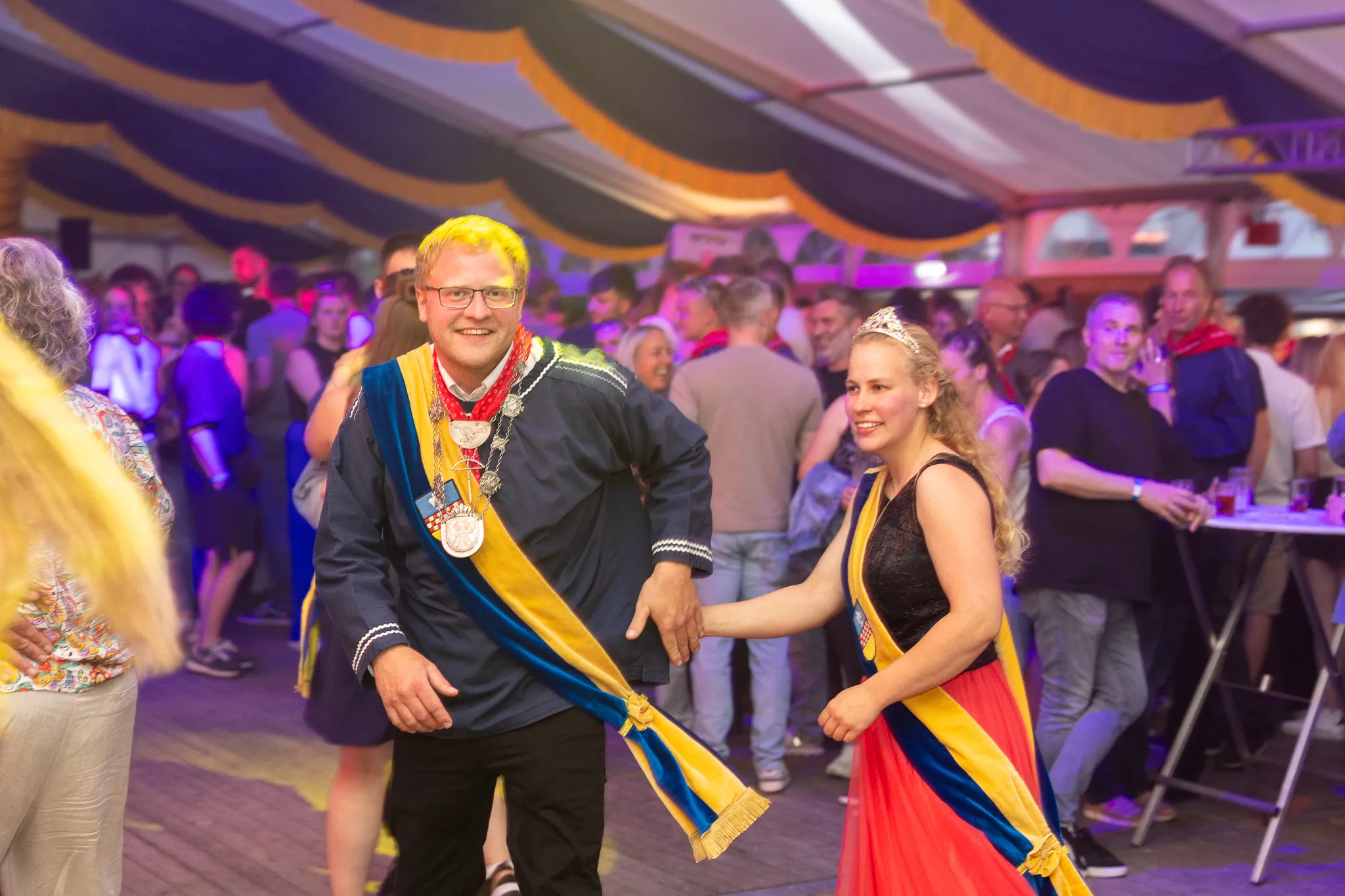 Ein Mann mit blonden Haaren und eine Frau mit einer Tiara tanzen im Festzelt, umgeben von feiernden Menschen.