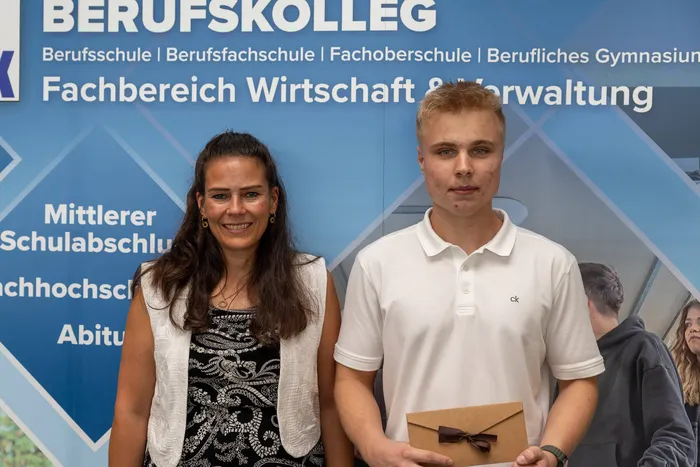 Zwei junge Erwachsene, eine Frau und ein Mann, stehen vor einer blauen Wand mit dem Aufdruck 