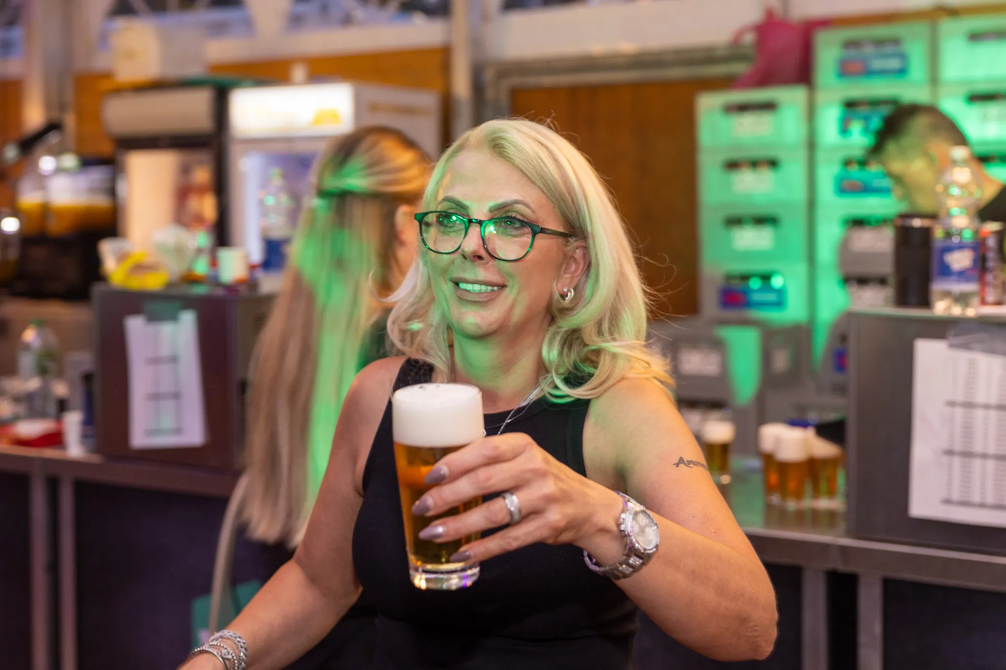 Eine blonde Frau mit Brille lächelt und hält ein Glas Bier in der Hand, im Hintergrund ein belebtes Festzelt.