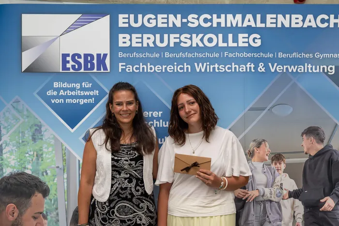 Zwei Frauen lächeln in die Kamera, während sie Urkunden halten. Im Hintergrund ist das Logo des Eugen-Schmalenbach Berufskollegs zu sehen.
