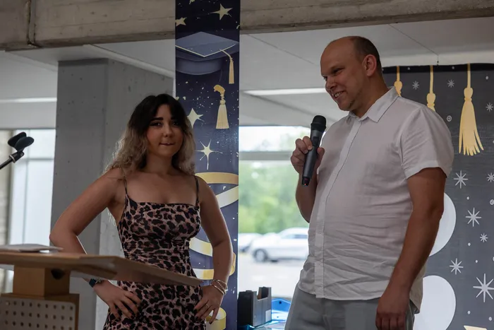 Ein Mann hält ein Mikrofon und spricht, während eine Frau neben ihm an einem Pult steht. Im Hintergrund ist ein Banner mit Abschlussmotiven zu sehen.