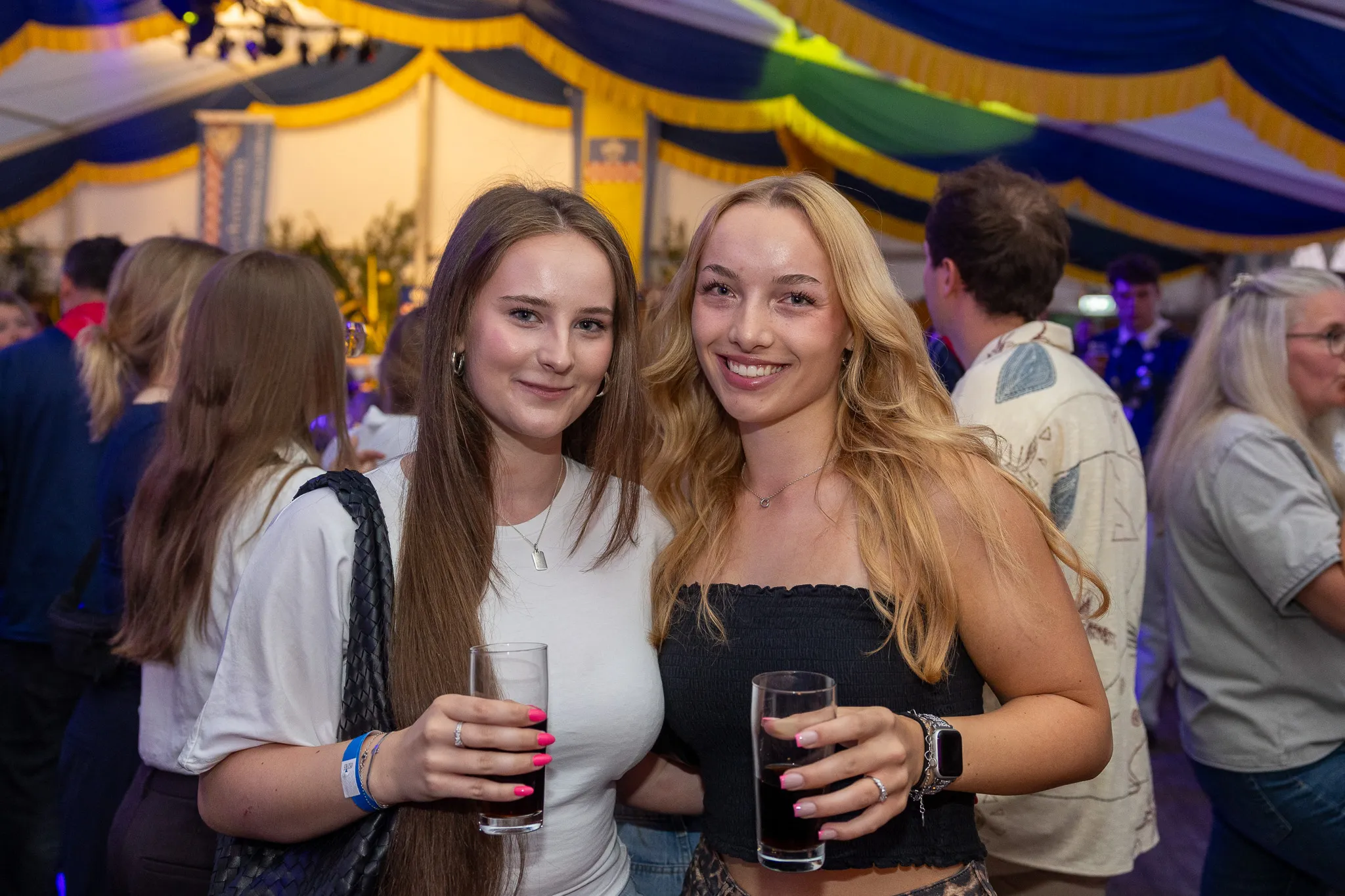 Zwei junge Frauen lächeln in die Kamera, jede hält ein Glas dunkle Flüssigkeit. Im Hintergrund sind weitere Menschen und die festliche Dekoration eines Zeltes zu sehen.