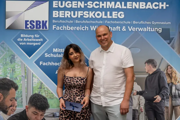 Ein Mann in einem weißen Hemd und eine junge Frau mit langen, dunklen Haaren stehen vor einem blauen Banner mit dem Aufdruck 