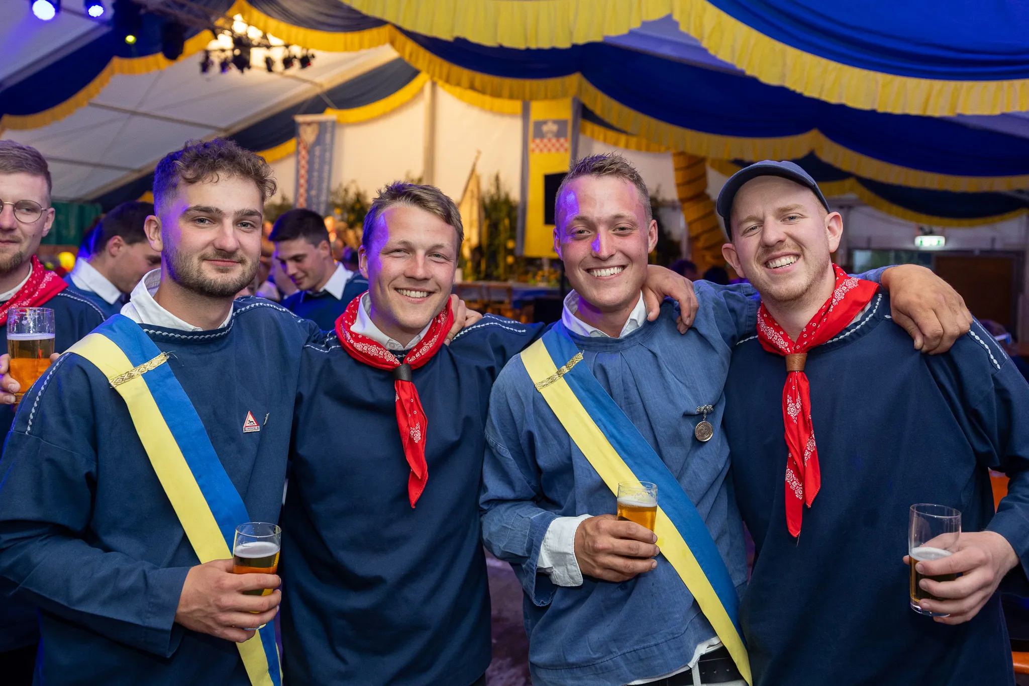 Vier junge Männer in traditioneller Tracht mit Bierkrügen stehen eng zusammen und lächeln in ein Zelt mit blauem Zeltdach und vielen Besuchern.