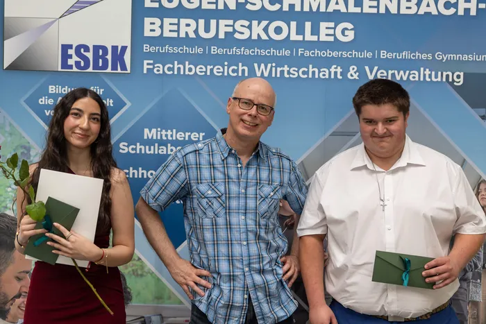 Drei Personen – eine junge Frau, ein Mann und ein junger Mann – stehen lächelnd vor einem Hintergrund mit dem Logo des ESBK (Eugen-Schmalenbach-Berufskolleg) und dem Schriftzug „Fachbereich Wirtschaft & Verwaltung“. Die Frau und der junge Mann halten jeweils eine grüne Schachtel, vermutlich eine Auszeichnung.