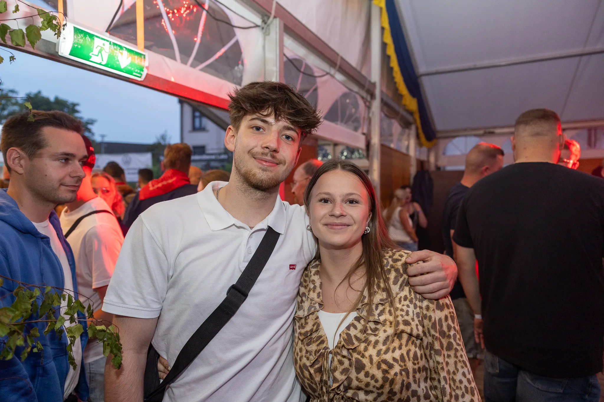 Ein junger Mann und eine junge Frau umarmen sich im überfüllten Festzelt des Bauernschützenballs. Im Hintergrund sind weitere Menschen und ein Notausgangsschild zu sehen.