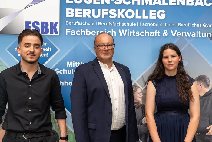 Drei Personen posieren vor einem Banner mit der Aufschrift „Berufskolleg“. Links ein junger Mann im schwarzen Hemd, in der Mitte ein Mann in einem beigen Anzug und rechts eine junge Frau mit langen braunen Haaren.