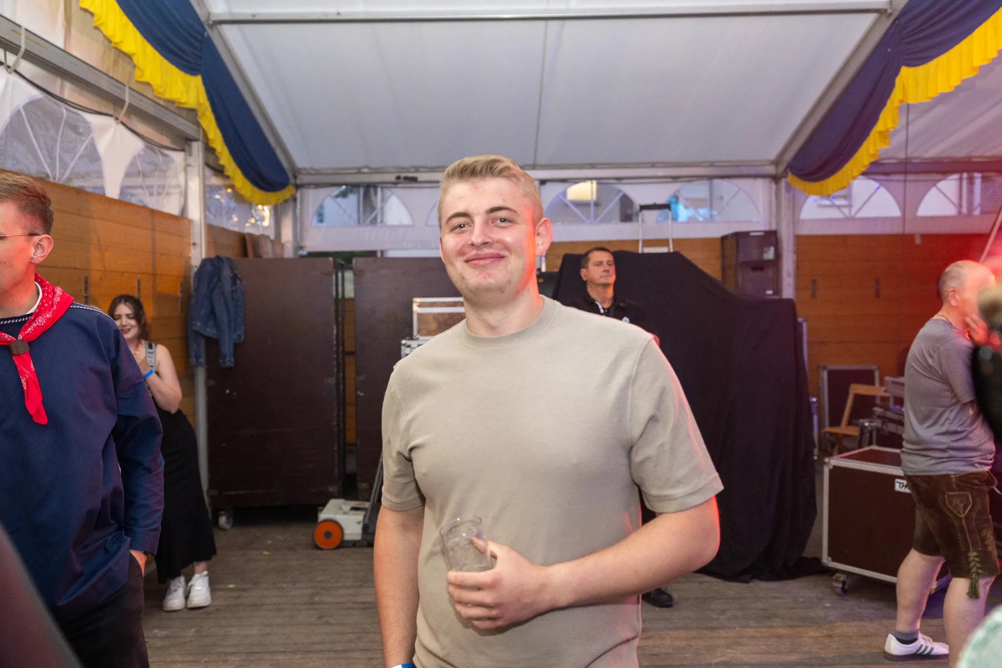 Ein junger Mann lächelt in einem Festzelt, im Hintergrund sind weitere Menschen und Bühnenelemente zu sehen.