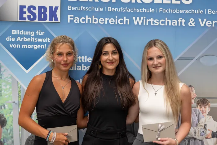 Drei junge Frauen stehen nebeneinander und lächeln in die Kamera. Im Hintergrund ein Banner der ESBK Berufsschule mit dem Hinweis auf den Fachbereich Wirtschaft.