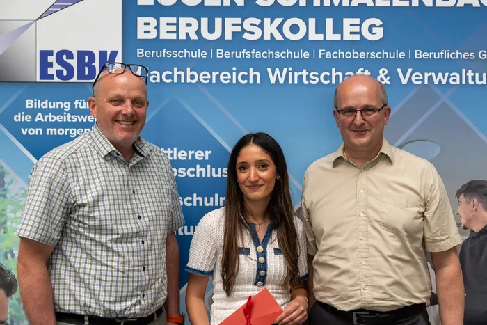Zwei Männer und eine junge Frau stehen vor einem Banner der ESBK Berufsschule. Die Frau hält eine rote Urkunde.