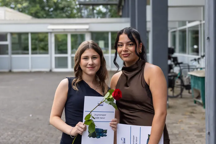 Zwei junge Frauen umarmen sich vor dem Gebäude der ESBK Halver und halten Zeugnisse sowie eine rote Rose. Im Hintergrund sind weitere Personen und Fahrräder zu sehen.