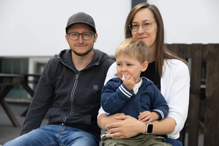 Ein Mann, eine Frau und ein kleines Kind sitzen nebeneinander und schauen in die Kamera. Das Kind isst etwas und wird von der Frau gehalten.