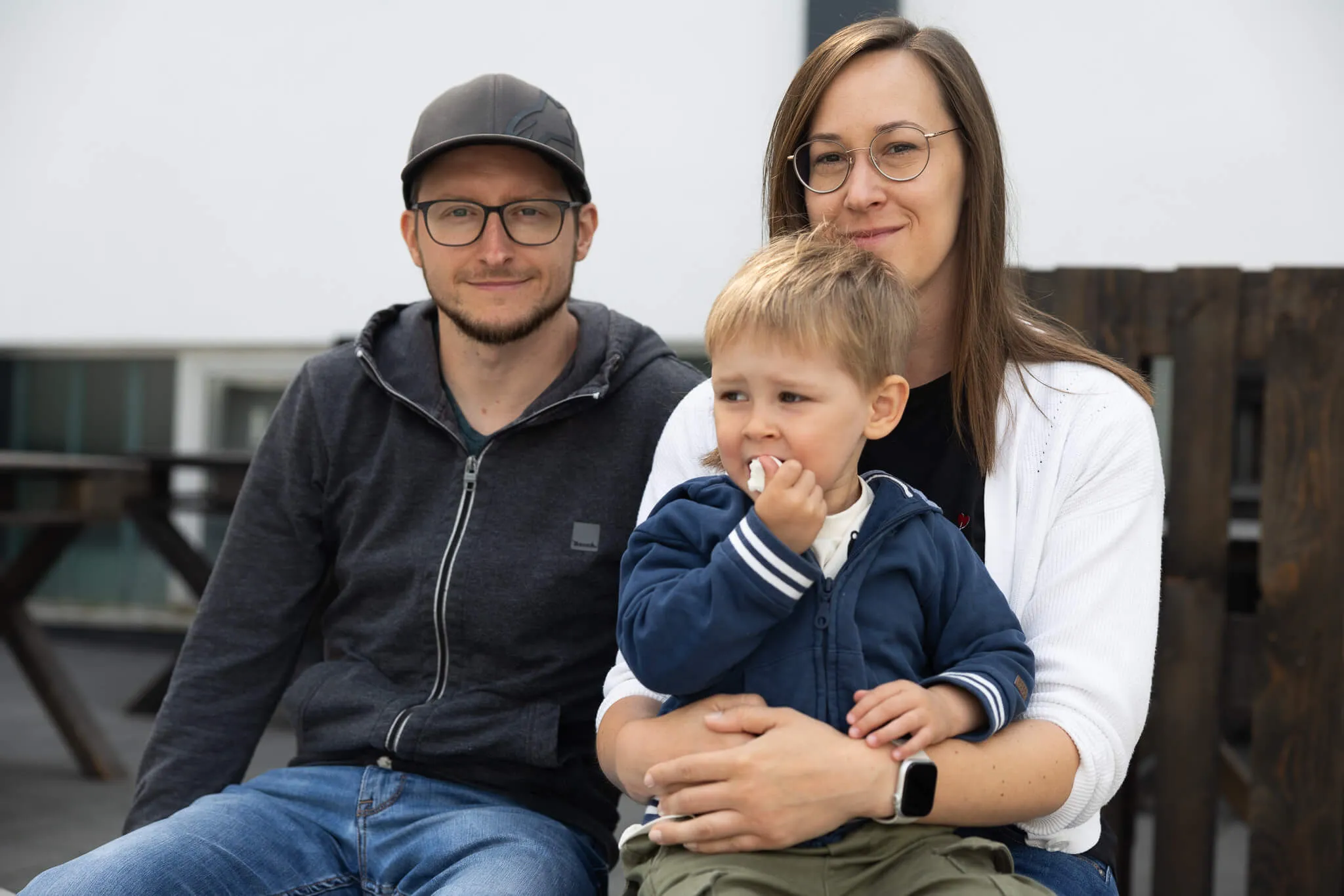 Ein Mann, eine Frau und ein kleines Kind sitzen nebeneinander und schauen in die Kamera. Das Kind isst etwas und wird von der Frau gehalten.