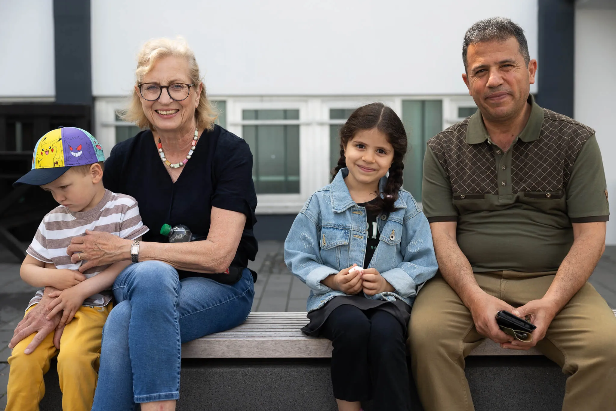 Eine ältere Frau, ein Mädchen und zwei Jungen sitzen nebeneinander auf einer Bank und lächeln in die Kamera. Im Hintergrund ist ein Gebäude zu sehen.