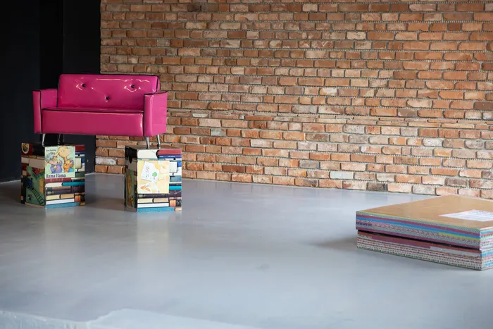 Ein kleines, pinkes Sofa steht vor einer Backsteinwand, flankiert von zwei Sockeln aus gestapelten Büchern. Rechts eine bunte Stapel von Büchern auf dem Boden.