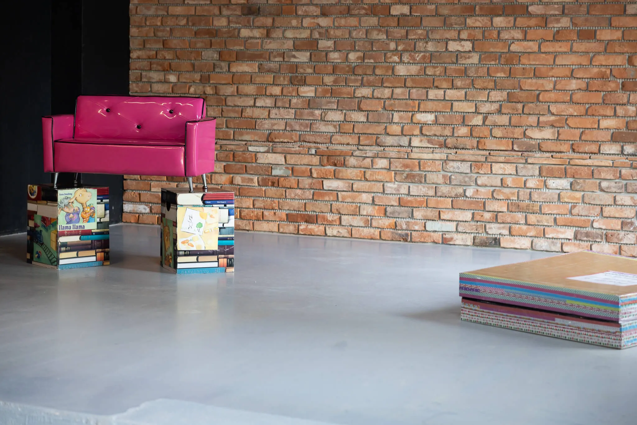 Ein kleines, pinkes Sofa steht vor einer Backsteinwand, flankiert von zwei Sockeln aus gestapelten Büchern. Rechts eine bunte Stapel von Büchern auf dem Boden.