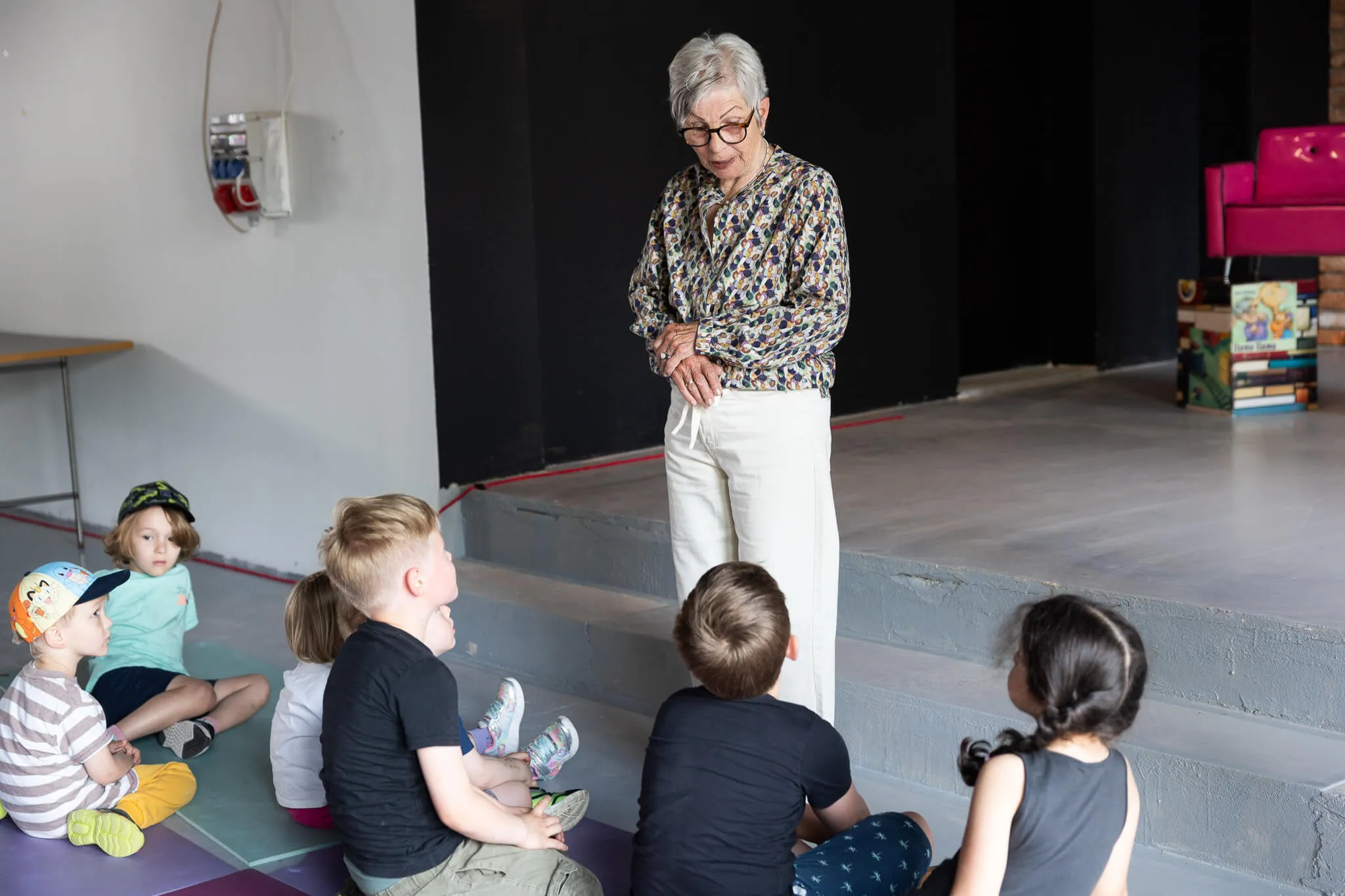 Eine ältere Frau steht vor einer Gruppe Kindergartenkinder und liest ihnen vor. Die Kinder sitzen auf Matten und schauen aufmerksam zu.