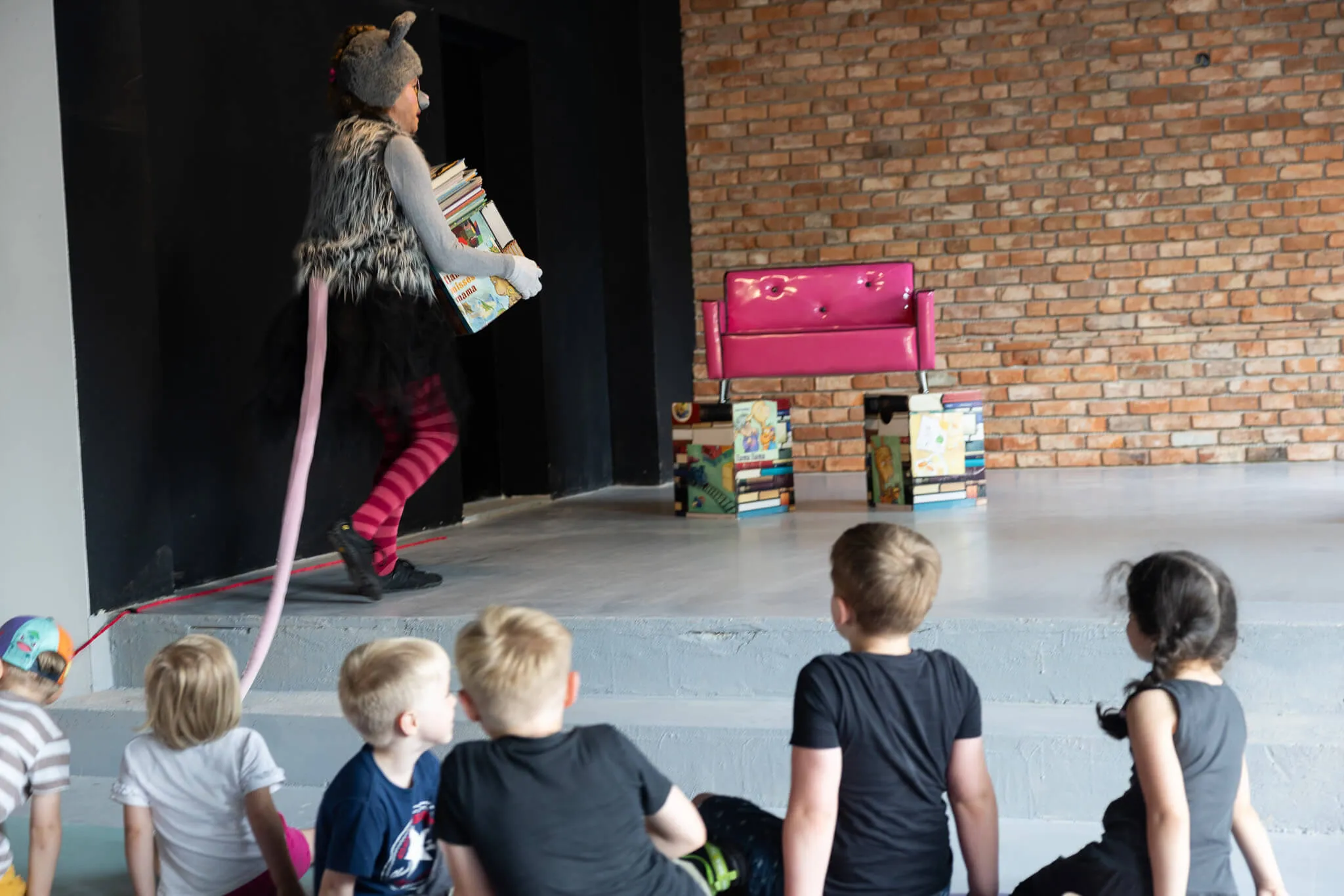 Eine Person in einem Mäusekostüm steht auf einer Bühne und hält einen Stapel Bücher, während eine Gruppe Kinder zusieht. Im Hintergrund steht ein pinkfarbenes Sofa und ein Bücherstapel.