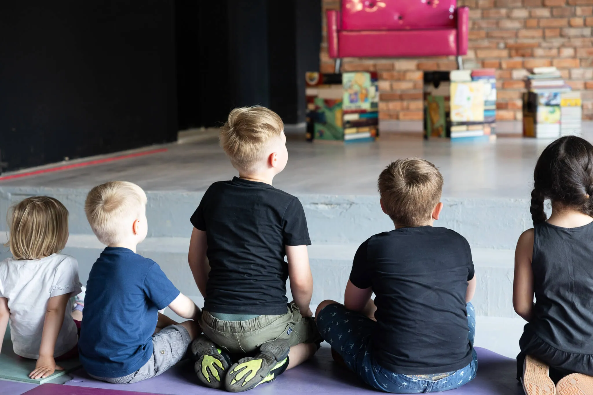 Fünf Kinder sitzen auf Matten mit dem Rücken zum Betrachter und schauen auf eine Person, die in einem Sessel sitzt und vor Bücherregalen steht.