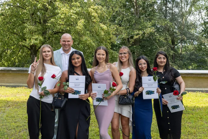 Gruppe von sechs jungen Frauen und einem Mann posiert im Freien, jeder hält eine rote Rose und eine Urkunde.