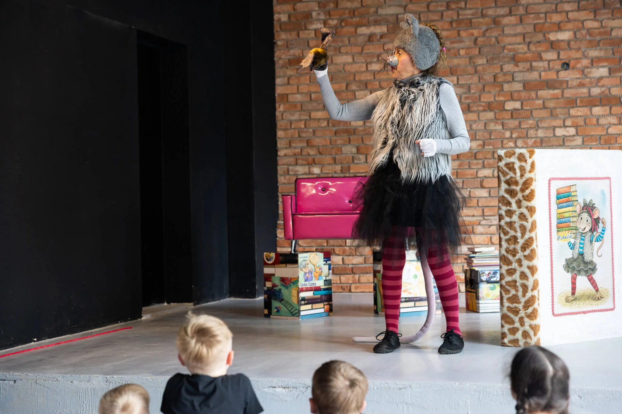 Person in Mäusekostüm steht auf einer Bühne und hält eine Stoffmaus, während Kinder im Publikum zusehen. Im Hintergrund sind Bücher und eine Illustration einer Maus zu sehen.