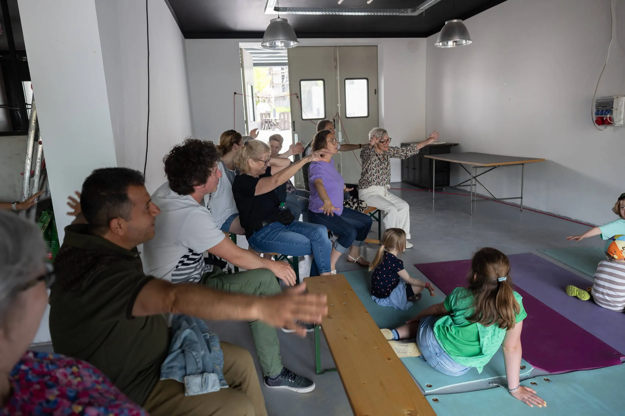 Gruppe von Erwachsenen und Kindern sitzt in einem hellen Raum und nimmt an einer interaktiven Aktivität teil, geleitet von einer Frau, die steht und gestikuliert.