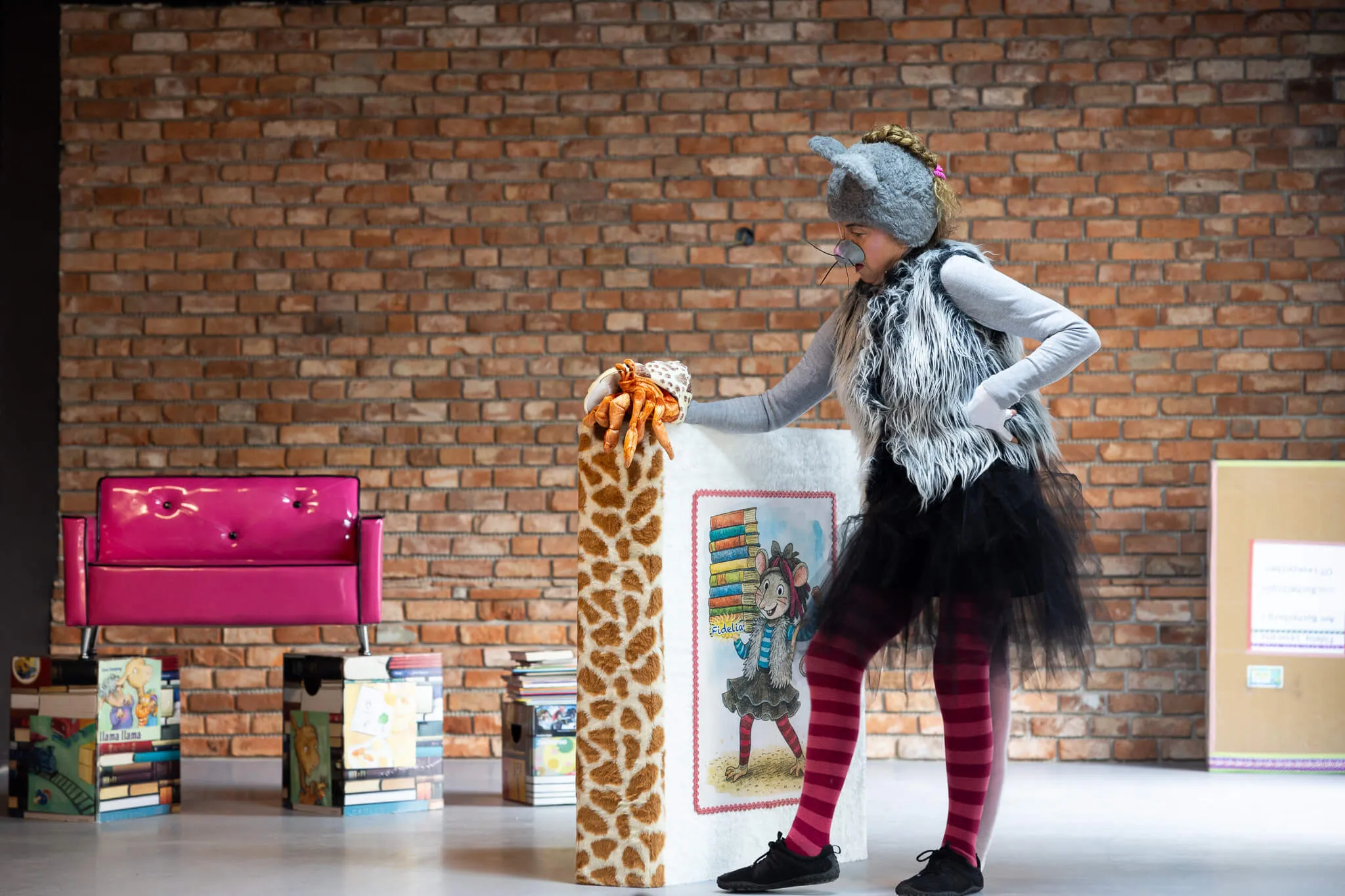 Ein Kind in einem Tierkostüm berührt eine Giraffenattrappe neben einem Bücherstapel mit einem Buchcover, das ein Mädchen mit Büchern zeigt. Im Hintergrund eine Backsteinwand und ein pinkfarbener Sessel.