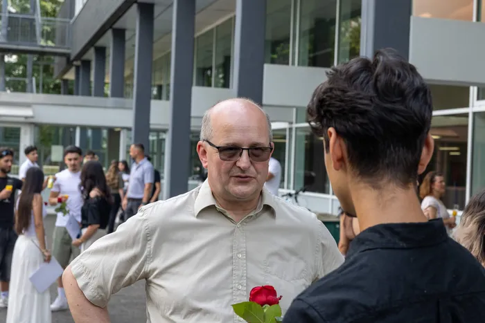 Ein Mann mit Brille und hellen Hemd unterhält sich mit einem jungen Mann mit dunklen, lockigen Haaren. Im Hintergrund sind weitere Personen und ein moderner Gebäudekomplex zu sehen.