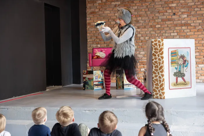 Person mit Mäusekostüm auf einer Bühne hält eine Handpuppe vor sich und spielt vor einer Gruppe von Kindergartenkindern.