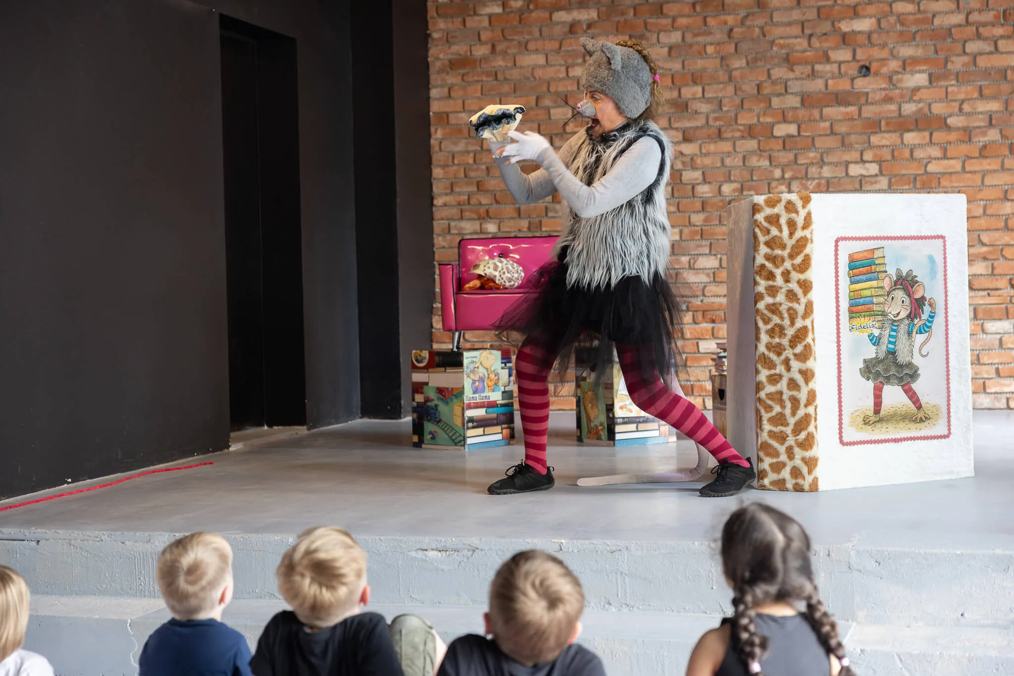 Person mit Mäusekostüm auf einer Bühne hält eine Handpuppe vor sich und spielt vor einer Gruppe von Kindergartenkindern.