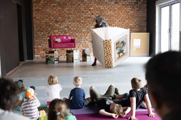 Eine Person mit einem Tierkostüm steht hinter einer selbstgebauten Kulisse und führt ein Puppentheater für eine Gruppe Kindergartenkinder auf.
