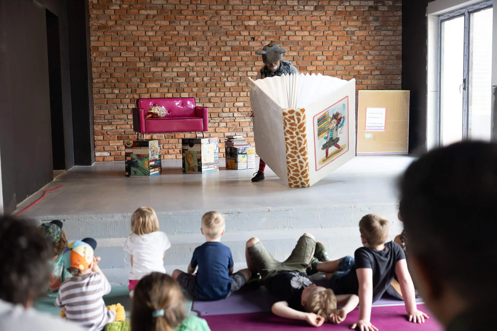 Eine Person mit einem Tierkostüm steht hinter einer selbstgebauten Kulisse und führt ein Puppentheater für eine Gruppe Kindergartenkinder auf.