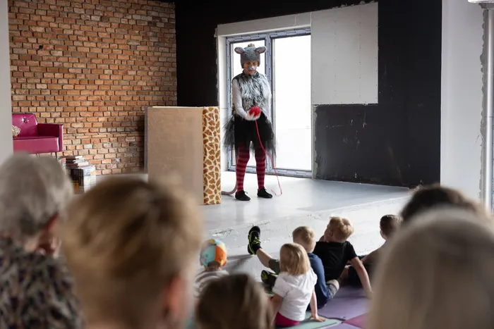 Person in Mäusekostüm steht auf einer Bühne vor Kindern, die auf Matten sitzen und zusehen. Im Hintergrund eine Backsteinwand und Fenster.