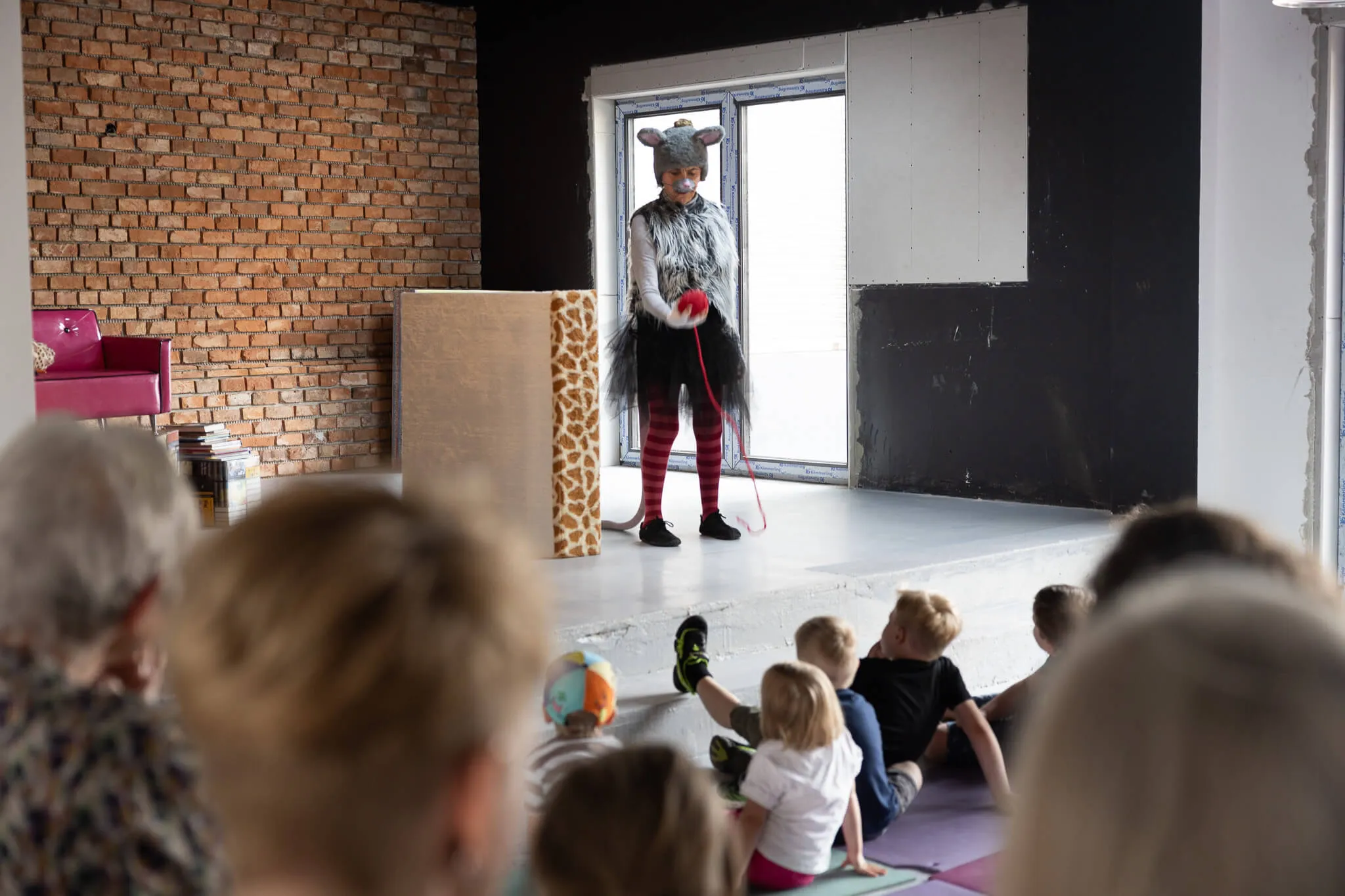 Person in Mäusekostüm steht auf einer Bühne vor Kindern, die auf Matten sitzen und zusehen. Im Hintergrund eine Backsteinwand und Fenster.