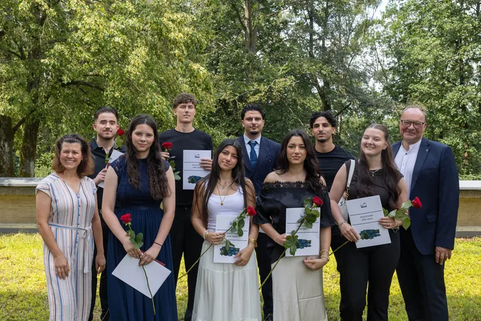 Gruppe von neun jungen Erwachsenen und zwei älteren Männern posiert im Freien vor Bäumen. Die Absolventen halten Urkunden und rote Rosen.