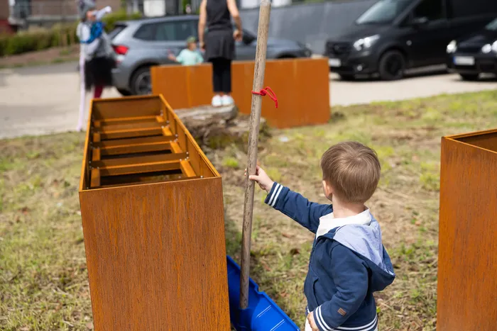 Ein kleiner Junge mit blauem Kapuzenpullover berührt einen langen Stab vor rostfarbenen, kastenförmigen Objekten im Freien. Im Hintergrund sind weitere Personen und geparkte Autos zu sehen.