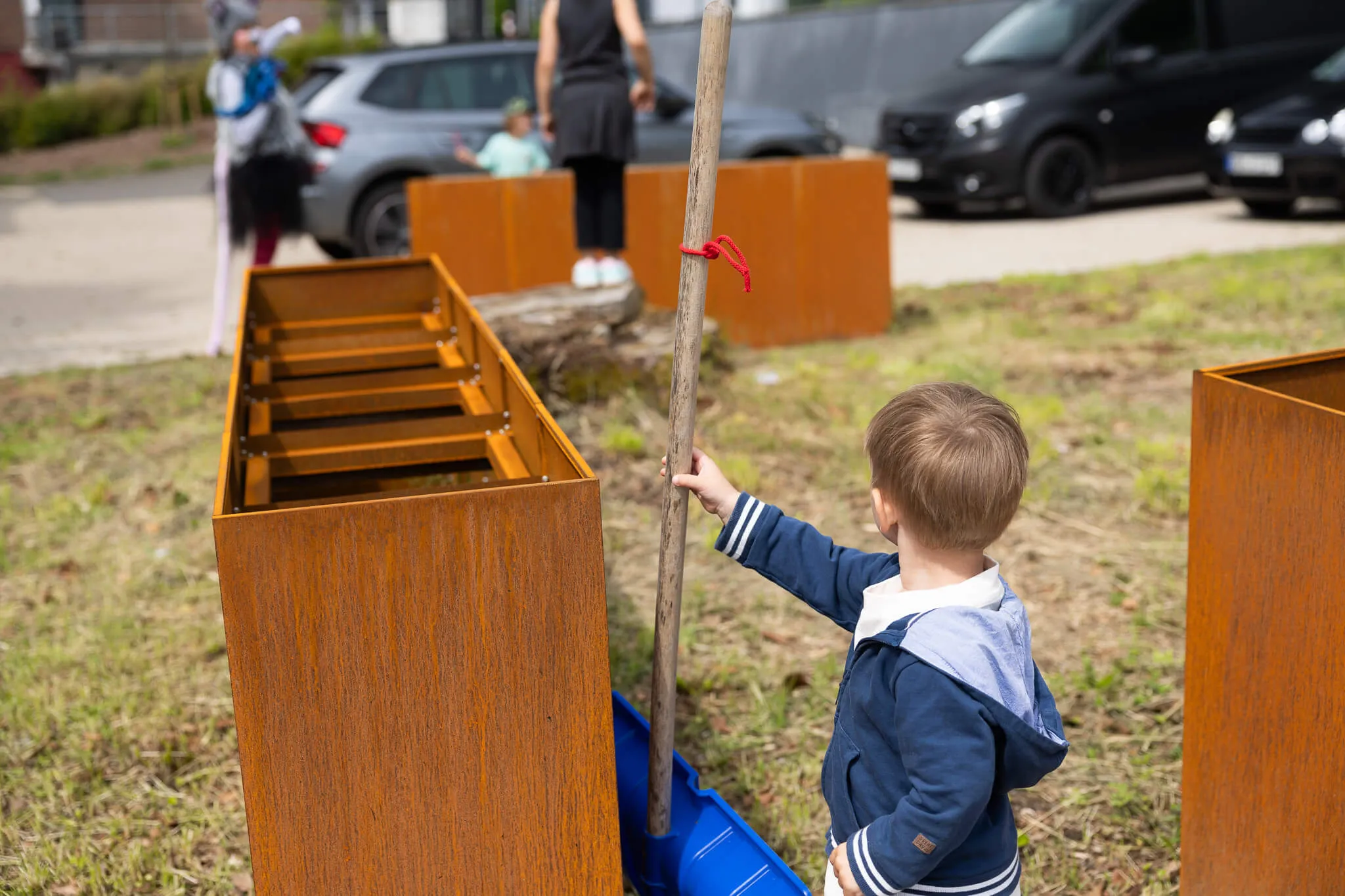 Ein kleiner Junge mit blauem Kapuzenpullover berührt einen langen Stab vor rostfarbenen, kastenförmigen Objekten im Freien. Im Hintergrund sind weitere Personen und geparkte Autos zu sehen.